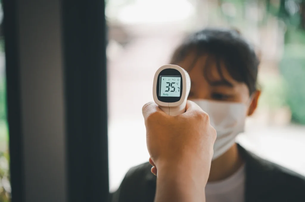 a temperature gun being held up to a lady with a face mask on. the temperature shows 35.7 degrees c