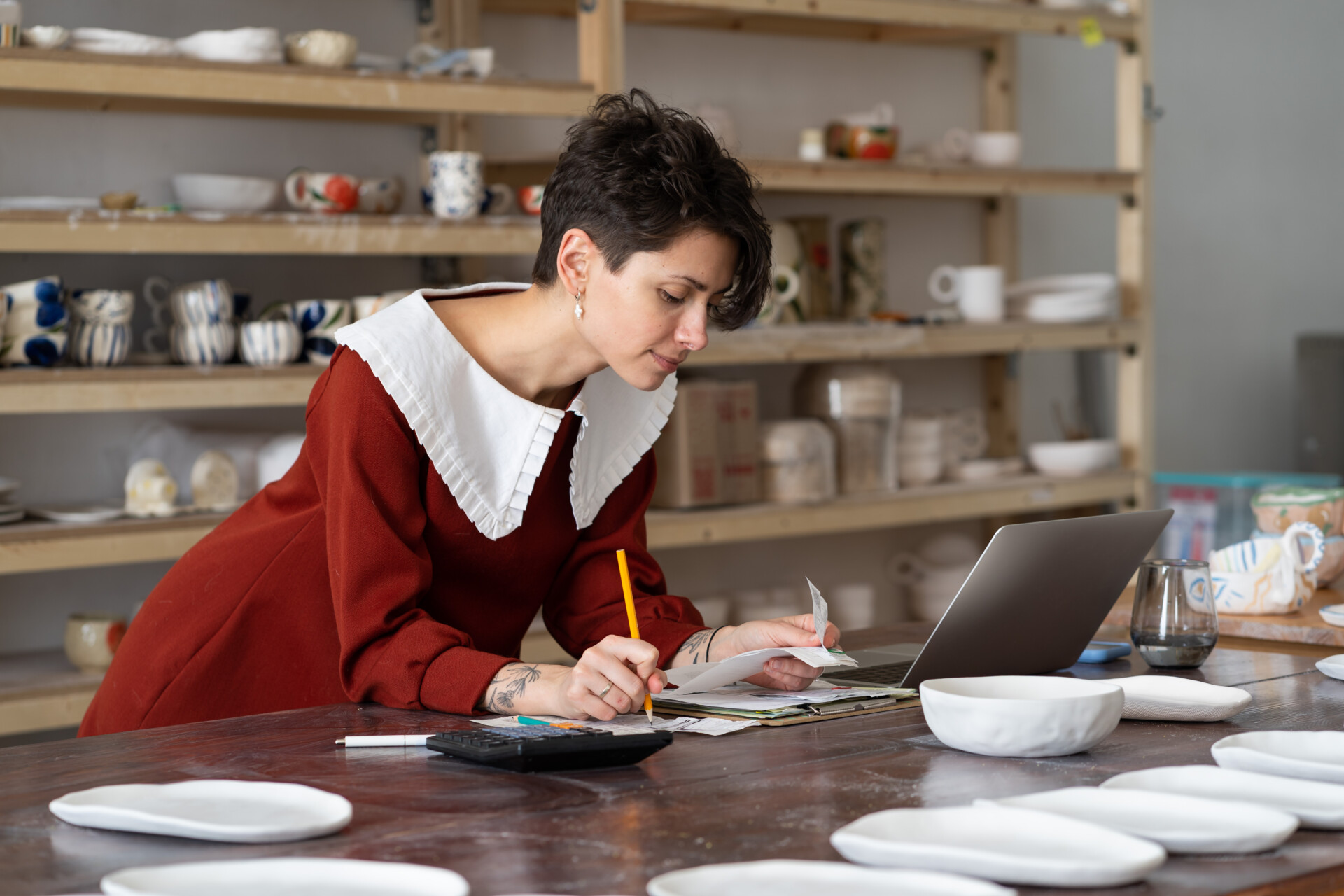 A small business owner completes accounts in a pottery studio using a laptop and calculator