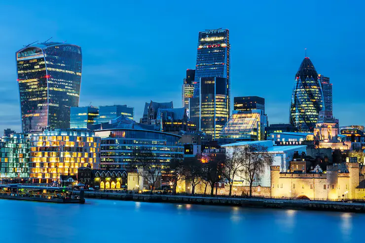 Illuminated London financial district skyline at dusk featuring prominent skyscrapers like the Walkie Talkie and The Gherkin along the River Thames.